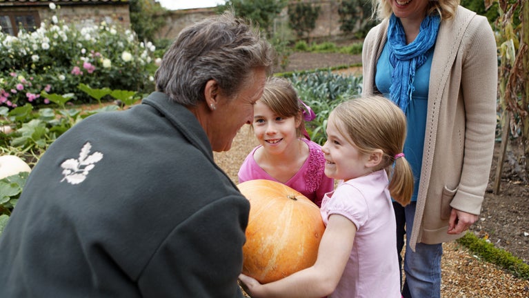 A woman and two children are in the kitchen garden holding a large pumpkin.